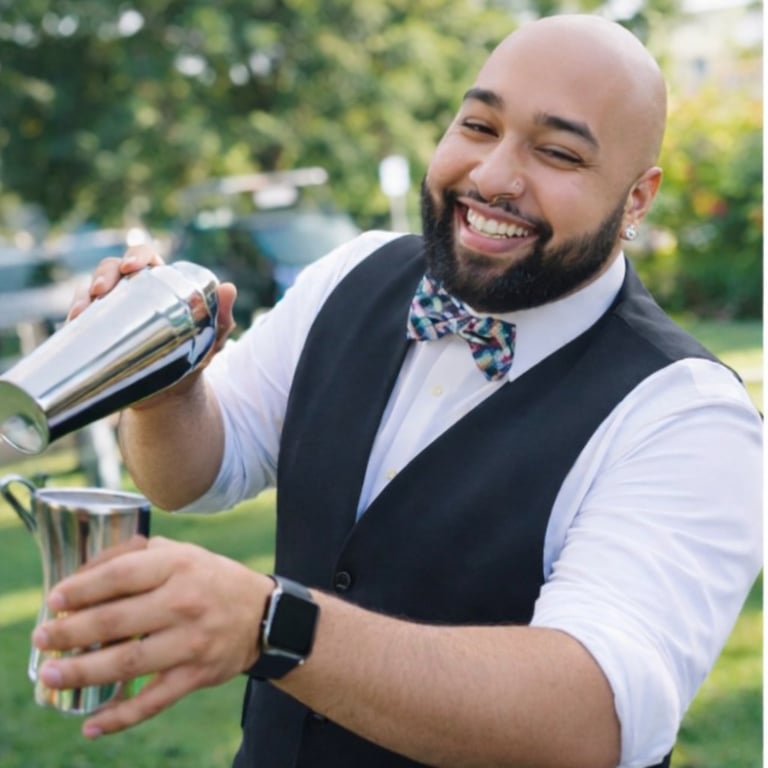 A smiling man in formal attire pouring a drink into a glass at an outdoor event, with green foliage in the background
