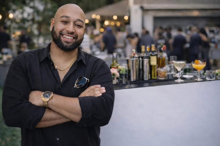 Smiling man in black shirt with arms crossed standing at outdoor bar with guests in background