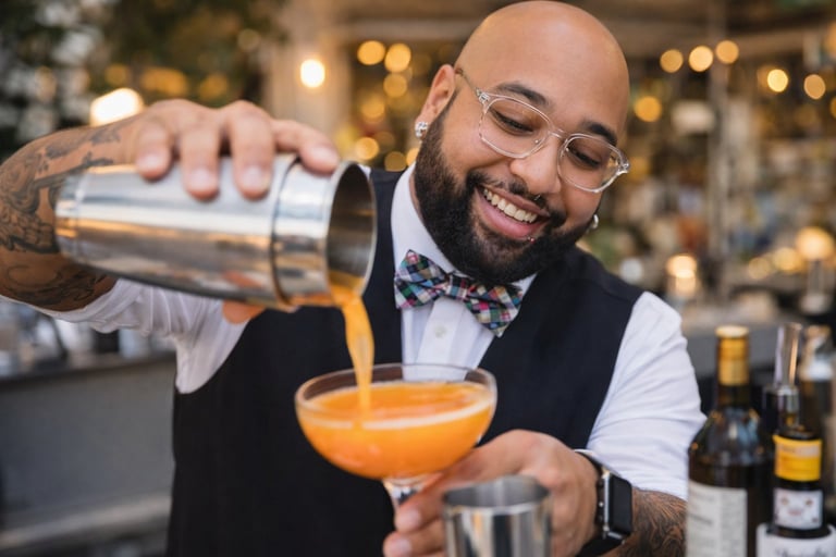 Bartender in vest and bow tie pouring an orange cocktail from a shaker into a glass at a bar with warm lighting